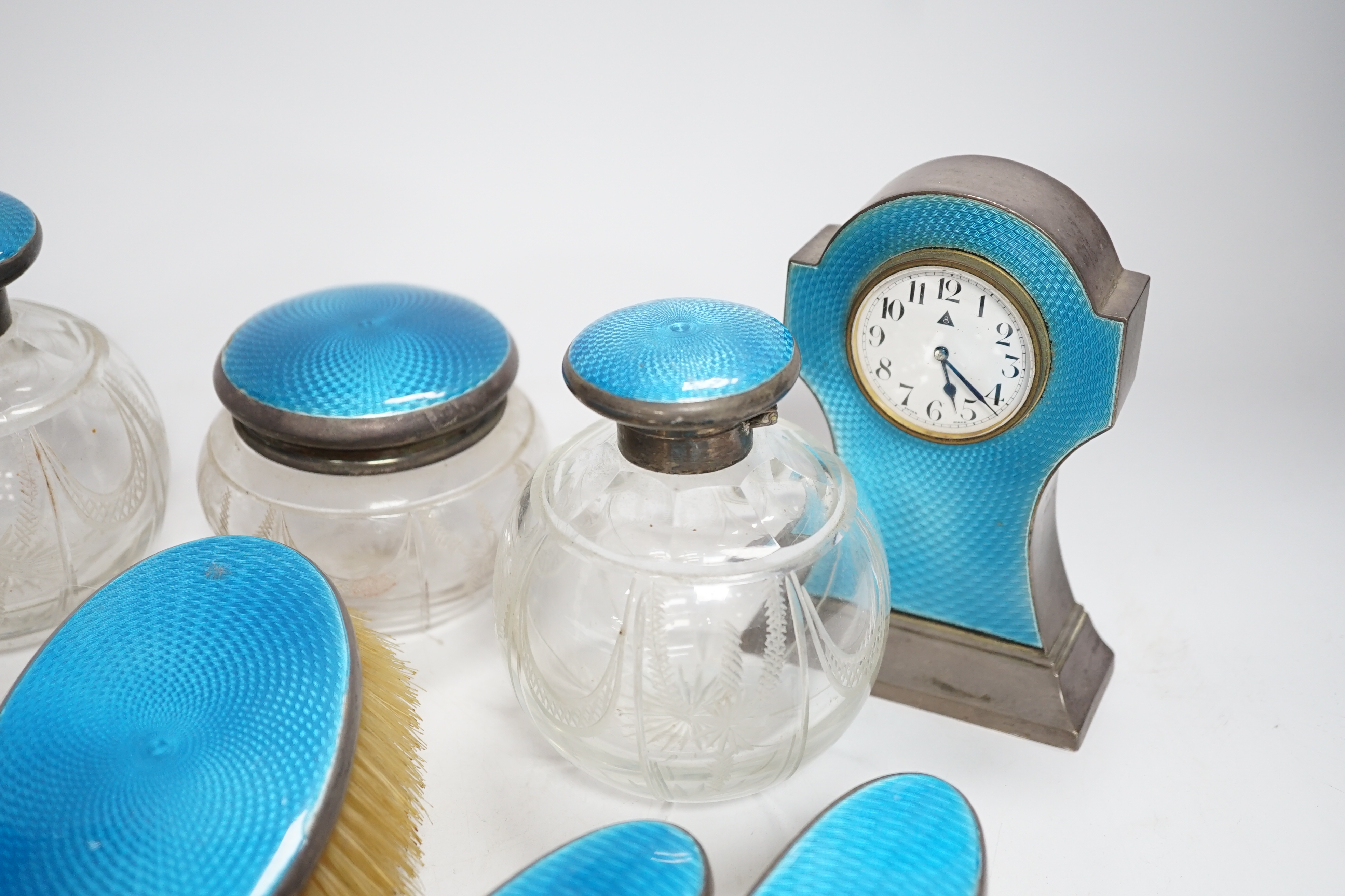 A George V silver and blue guilloche enamelled ten piece dressing table set, comprising four brushes, a hand mirror, two mounted glass toilet jars, two mounted glass scent and a mantel timepiece, 12.8cm, C.S Green & Co,